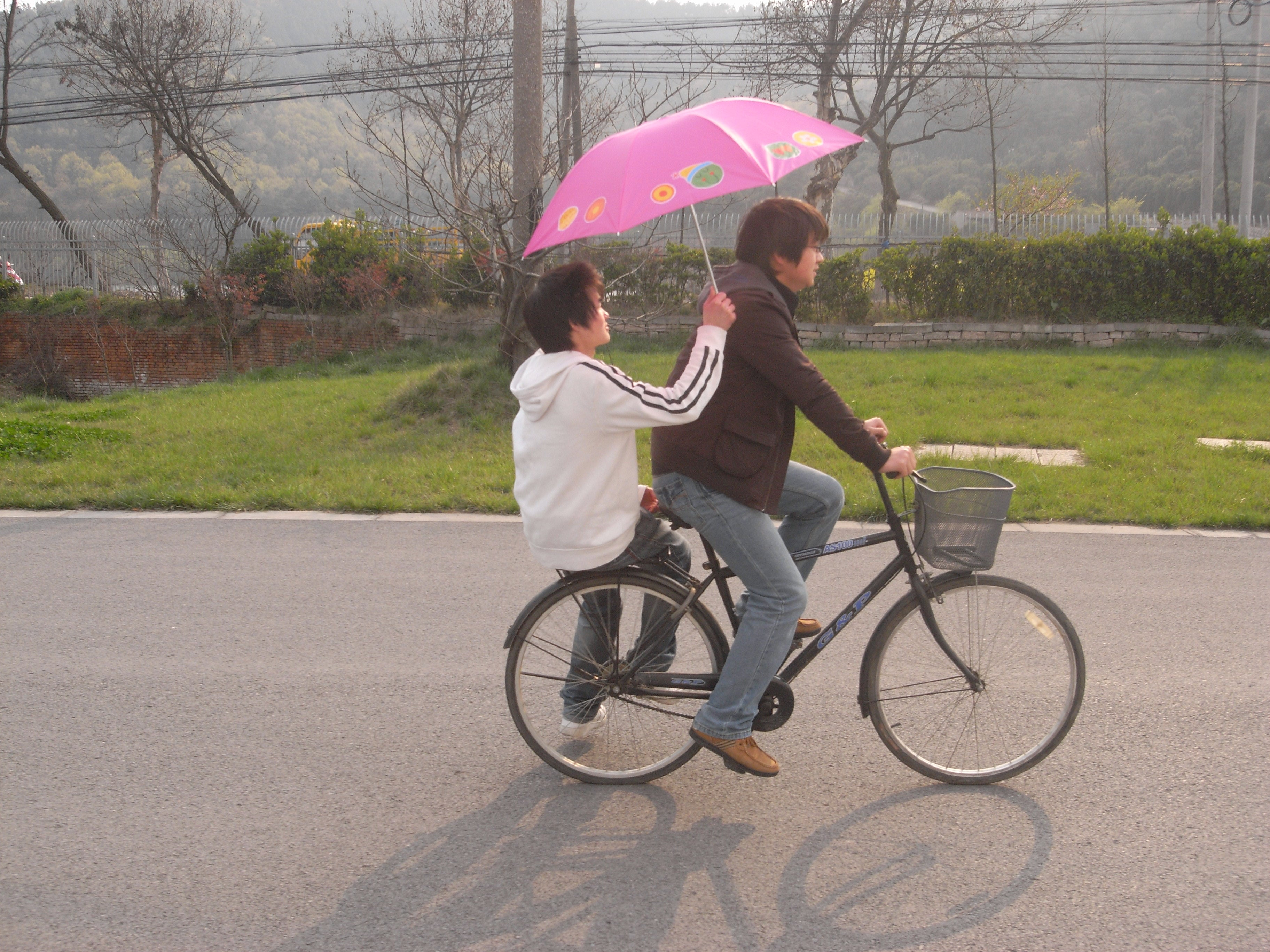 Two people riding a bicycle on a road with a pink umbrella
